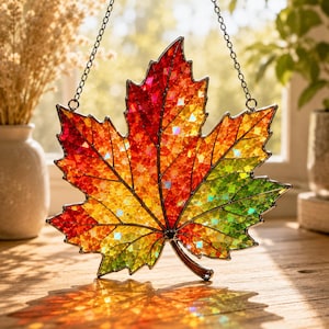 May include: A stained glass maple leaf suncatcher featuring a vibrant gradient of red, orange, yellow, and green iridescent glass segments held together by dark metal cames. The leaf hangs from a thin silver chain, casting colorful light reflections.