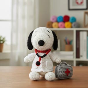 May include: A crocheted Snoopy doll dressed as a doctor, wearing a white coat and a red and black stethoscope. A small gray crocheted medical bag with a red cross sits beside the doll. The doll has black ears and a black nose.