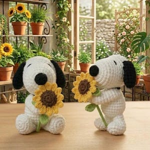 May include: Two crochet Snoopy figures, one sitting and one standing, each holding a yellow and brown sunflower. The figures are white with black ears and a black nose. The background includes potted plants and a window.