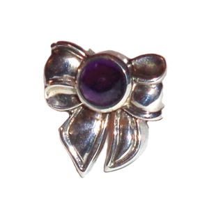 May include: A silver-toned bow-shaped ring with a round, deep purple gemstone at its center. The ring's design features detailed bow loops and flowing ribbon ends, creating a delicate and elegant appearance. The gemstone is a focal point.