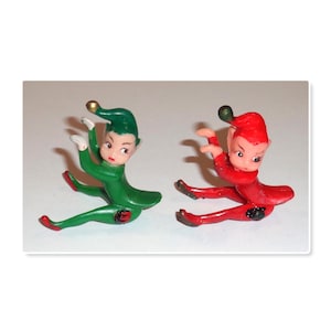 May include: Two small elf figurines in a seated position, one dressed in a solid green suit with a gold-tipped hat and the other in a solid red suit with a dark-tipped hat. Both figures have white gloves, pointed ears, and black circular accents.
