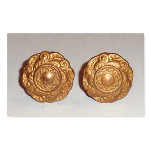 May include: Two antique gold-colored cabinet knobs. Each knob has a circular design with a central raised circle and ornate, swirling patterns around the edge. The knobs are made of metal and have a slightly aged appearance.