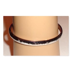 May include: A silver-colored bangle bracelet with a repeating star and leaf pattern. The bracelet is round and appears to be made of metal. The bracelet is worn on a mannequin arm.