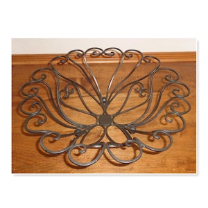 May include: A decorative, dark metal bowl with a floral design. The bowl features a central circular base with radiating curved lines and decorative swirls. The bowl is designed to hold fruit or other decorative items.