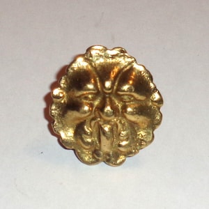May include: A gold-colored, round button with a detailed face design. The button features a stylized face with prominent features and a textured surface, likely for decorative purposes. The button is likely made of metal.