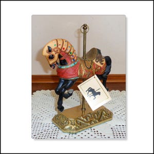 May include: A decorative carousel horse figurine with a red, gold, and black color scheme. The horse has a detailed saddle and is adorned with jewels. The base is gold-colored with ornate detailing, and a tag with a horse silhouette hangs from the pole.