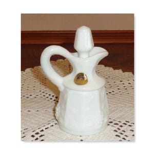 May include: A white milk glass cruet featuring a textured grape and leaf pattern on the body, a curved handle, a pointed stopper, and a small circular gold foil label on the neck. The vessel has a wide base and a narrow spout for pouring.