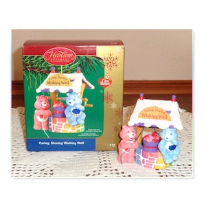 May include: A Care Bears Heirloom Collection ornament featuring a wishing well with a pink and blue bear. The well has a white roof with the words "Caring, Sharing Wishing Well". The box is visible in the background.