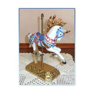 May include: A white carousel horse figurine with a gold mane and tail, adorned with a blue saddle and colorful jewels. The horse is mounted on a gold-colored base with an ornate design, and a gold pole. A tag hangs from the base.