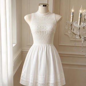 Pode incluir: Um vestido branco sem mangas com gola alta e corpete justo com padrões de malha intrincados. A cintura é marcada, transicionando para uma saia rodada com faixas horizontais e um detalhe delicado de ponto aberto na bainha inferior.