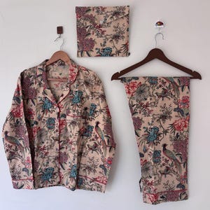 Könnte beinhalten: Ein beiges Pyjama-Set mit Blumen- und Vogelmuster, bestehend aus einem Hemd mit Knöpfen, einer Hose und einem passenden Aufbewahrungsbeutel. Das Set hat rote und blaue Akzente und wird auf Holzbügeln präsentiert.