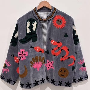 Puede incluir: Chaqueta de terciopelo gris con dise&ntilde;os bordados coloridos. La chaqueta presenta una bota de vaquero, una serpiente roja, un sol, flores y otras formas caprichosas. La chaqueta est&aacute; colgada de una percha de madera.