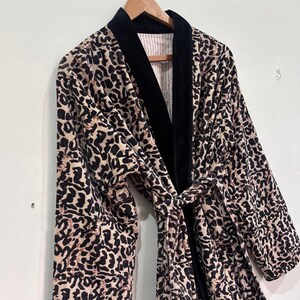 May include: A leopard print robe with a black velvet collar and tie. The robe is hanging on a wooden hanger against a white wall. The interior lining has a pink and white striped pattern.