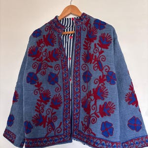 Puede incluir: Chaqueta azul con bordados florales rojos y azules. La chaqueta tiene un forro a rayas y está colgada de una percha de madera. El bordado es detallado y cubre la parte delantera y las mangas de la chaqueta.