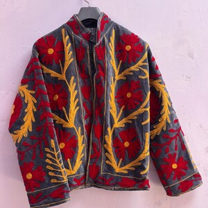 Puede incluir: Chaqueta gris con cuello alto y bordados detallados. La chaqueta presenta diseños florales rojos y patrones de hojas amarillas. El bordado crea una apariencia texturizada. La chaqueta está colgada en una percha negra.