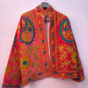 Puede incluir: Chaqueta color coral con intrincados bordados en turquesa, amarillo y naranja. La chaqueta presenta un cuello mandarín y un forro a rayas. Los bordados incluyen diseños florales y de cachemira.