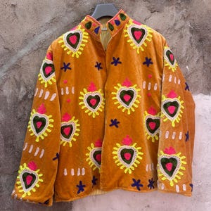May include: A mustard yellow velvet jacket featuring embroidered heart motifs in bright pink, green, and yellow. The jacket has a high collar, long sleeves, and scattered small blue star and white dash embroidery across the front and sleeves.
