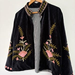 May include: A black velvet jacket featuring a mandarin collar and long sleeves. The jacket is adorned with intricate floral embroidery in shades of pink and olive green. The interior lining displays a black and white vertical striped pattern.