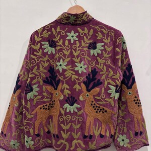 May include: A purple jacket with intricate embroidery featuring deer and floral patterns. The jacket has long sleeves and a high collar. The embroidery includes shades of green, brown, and navy blue, creating a detailed and artistic design.