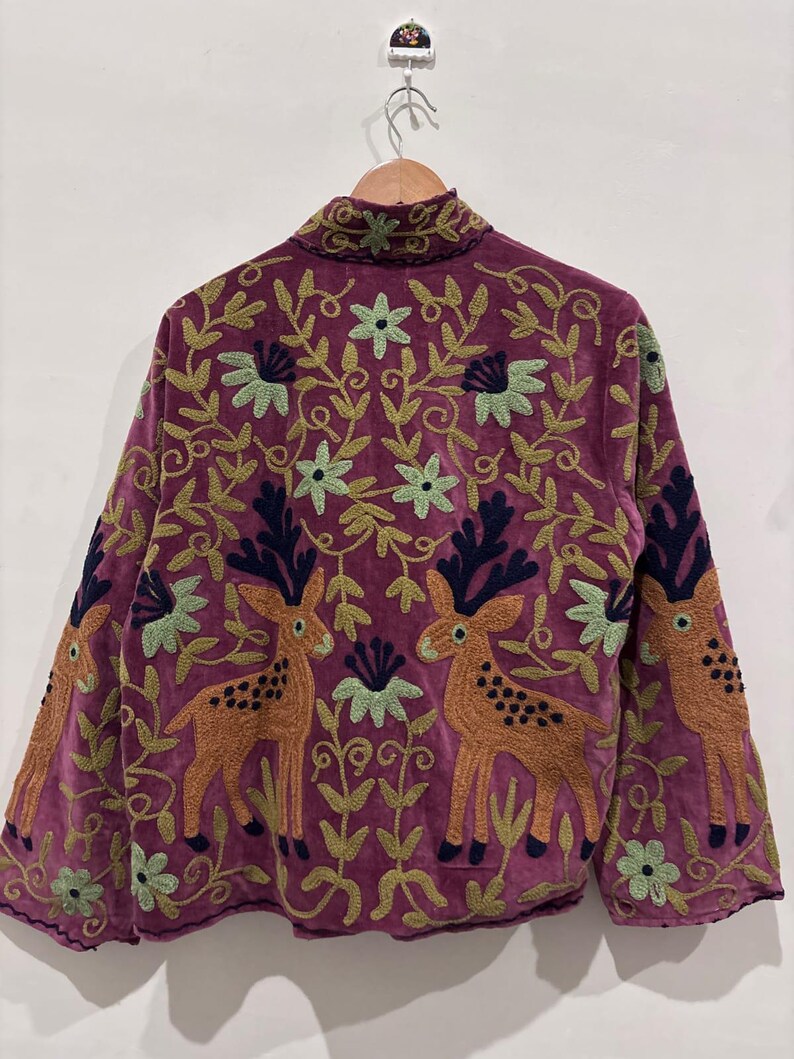 May include: A purple jacket with intricate embroidery featuring deer and floral patterns. The jacket has long sleeves and a high collar. The embroidery includes shades of green, brown, and navy blue, creating a detailed and artistic design.