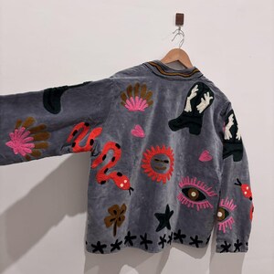 Puede incluir: Chaqueta de terciopelo gris con dise&ntilde;os bordados coloridos. La chaqueta presenta una serpiente, un sol, botas, ojos y patrones florales. La chaqueta est&aacute; colgada en una percha de madera contra una pared blanca.
