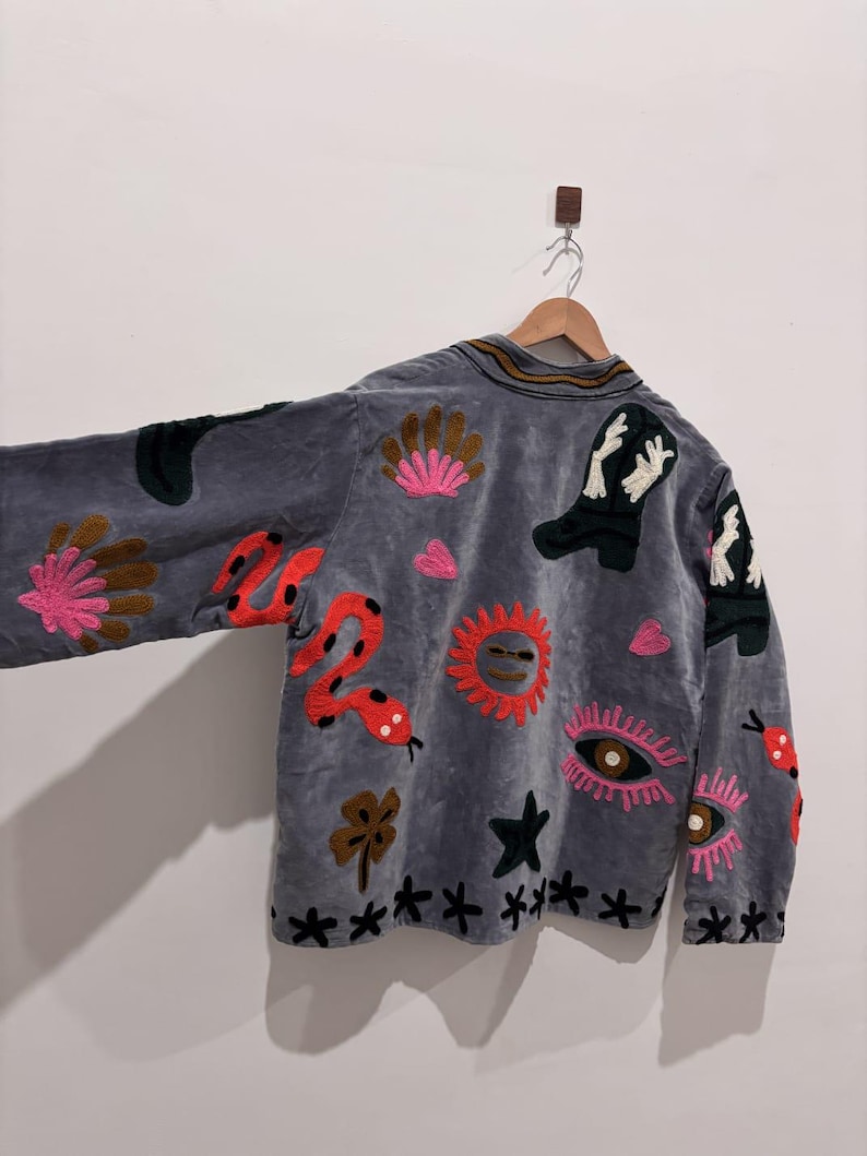 Puede incluir: Chaqueta de terciopelo gris con dise&ntilde;os bordados coloridos. La chaqueta presenta una serpiente, un sol, botas, ojos y patrones florales. La chaqueta est&aacute; colgada en una percha de madera contra una pared blanca.