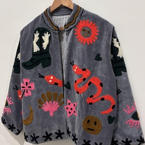Puede incluir: Chaqueta gris con dise&ntilde;os bordados coloridos, incluyendo una serpiente roja, un sol y una bota de vaquero. La chaqueta presenta un patr&oacute;n floral y de estrellas negras en los bordes y mangas. Colgada en una percha de madera.