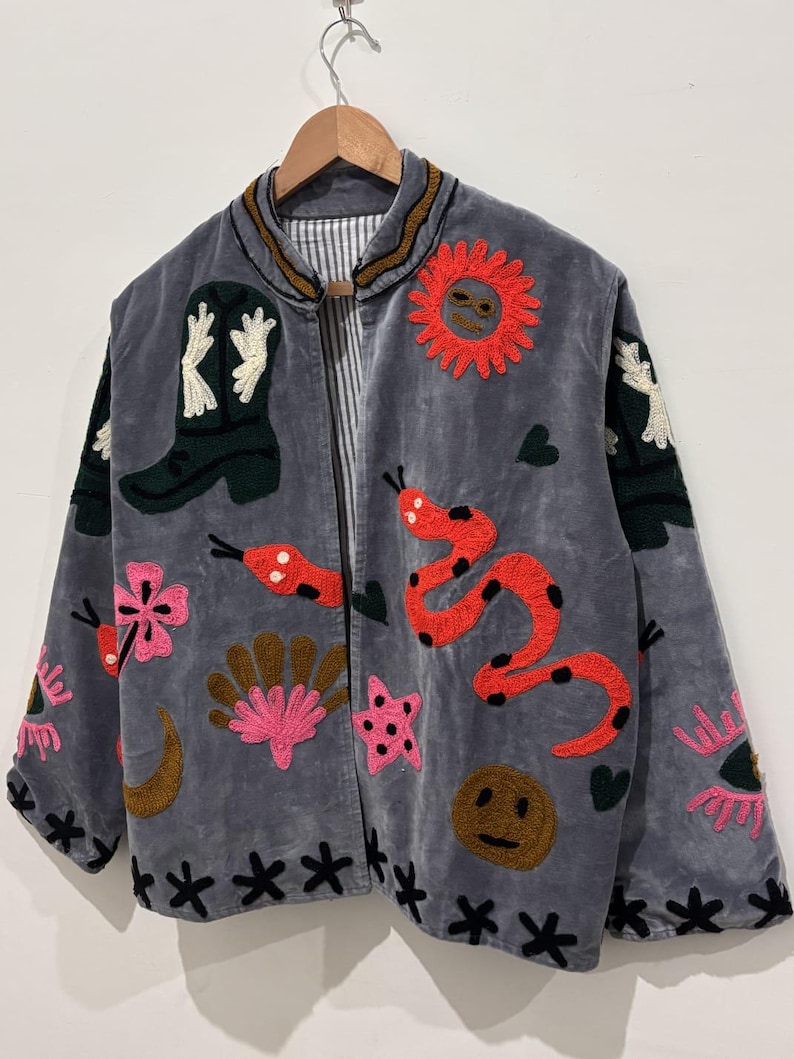 Puede incluir: Chaqueta gris con dise&ntilde;os bordados coloridos, incluyendo una serpiente roja, un sol y una bota de vaquero. La chaqueta presenta un patr&oacute;n floral y de estrellas negras en los bordes y mangas. Colgada en una percha de madera.