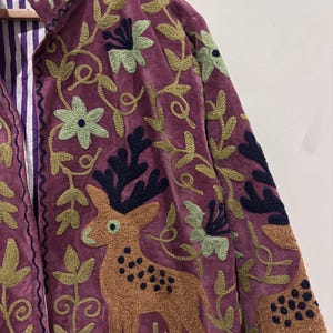May include: A burgundy velvet jacket with intricate embroidery. The jacket features a deer and floral design in shades of brown, green, and navy. The interior lining is striped with purple and white.