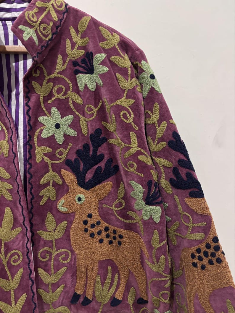May include: A burgundy velvet jacket with intricate embroidery. The jacket features a deer and floral design in shades of brown, green, and navy. The interior lining is striped with purple and white.