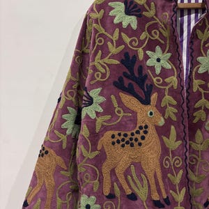 May include: A plum-colored jacket with intricate embroidery featuring deer, flowers, and foliage in shades of brown, green, and navy. The jacket's design includes a collar and a striped lining, showcasing a blend of textures and patterns.