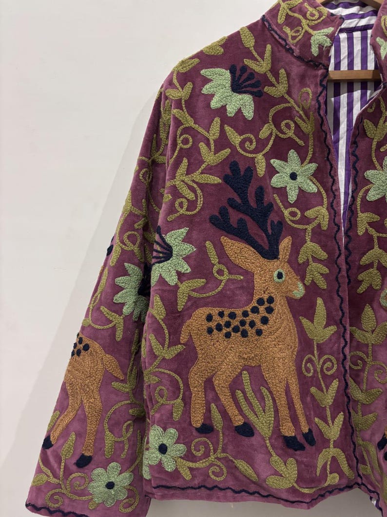 May include: A plum-colored jacket with intricate embroidery featuring deer, flowers, and foliage in shades of brown, green, and navy. The jacket's design includes a collar and a striped lining, showcasing a blend of textures and patterns.