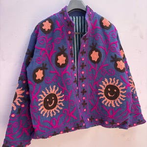 Könnte beinhalten: Eine blaue Jacke mit leuchtend pinkfarbenen Stickereien in Form von Ranken und dunkelbraunen kreisförmigen Motiven. Das Design zeigt stilisierte Sonnen mit lächelnden Gesichtern in Dunkelbraun und Pink sowie granatapfelförmige Akzente. Gestreiftes Innenfutter.