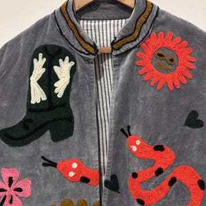 Puede incluir: Chaqueta de terciopelo gris con detalles bordados. Presenta una bota de vaquero verde, serpientes rojas, un sol y dise&ntilde;os florales. El cuello est&aacute; adornado con costuras doradas y marrones. La chaqueta est&aacute; colgada en una percha de madera.