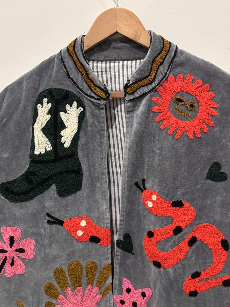 Puede incluir: Chaqueta de terciopelo gris con detalles bordados. Presenta una bota de vaquero verde, serpientes rojas, un sol y dise&ntilde;os florales. El cuello est&aacute; adornado con costuras doradas y marrones. La chaqueta est&aacute; colgada en una percha de madera.