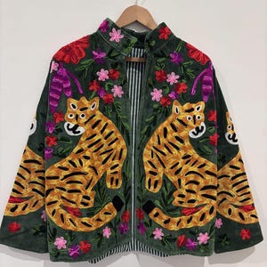 Puede incluir: Chaqueta verde oscuro con bordados de tigres y detalles florales. La chaqueta tiene un cuello alto, una cremallera negra y un forro a rayas. Los tigres son amarillos y negros, y las flores son rosas, rojas y moradas.