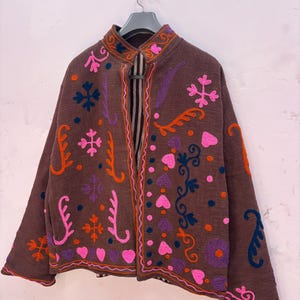 Puede incluir: Una chaqueta marrón con cuello mao y mangas largas, decorada con bordados vibrantes y multicolores en patrones de corazones, flores y formas abstractas en rosa, naranja, morado y azul oscuro, colgada en una percha gris.