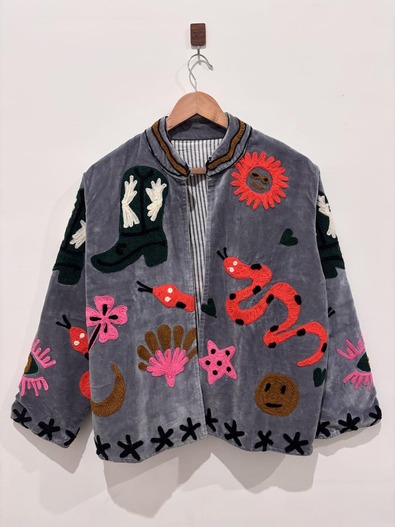 Puede incluir: Chaqueta de terciopelo gris con dise&ntilde;os bordados coloridos. La chaqueta presenta una bota de vaquero, una serpiente roja, un sol, flores y otras formas caprichosas. La chaqueta est&aacute; colgada de una percha de madera.