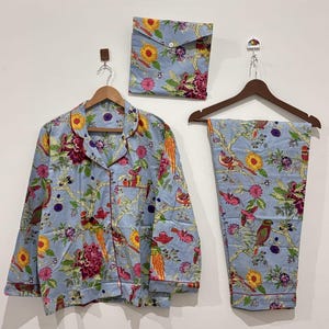 Könnte beinhalten: Ein hellblaues Pyjama-Set mit Blumen- und Vogelmuster. Das Set beinhaltet ein Hemd mit Knöpfen, eine Hose und eine passende Tasche. Der Druck zeigt rote, gelbe und grüne Blumen und Vögel. Hemd und Hose hängen an Holzbügeln.