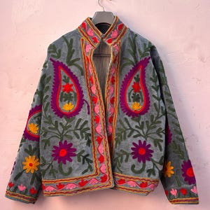 Puede incluir: Chaqueta gris con intrincados bordados florales en tonos morados, rojos, rosas y amarillos. La chaqueta presenta un cuello alto y está adornada con diseños en forma de cachemira y patrones de hojas. La chaqueta está colgada en una percha.