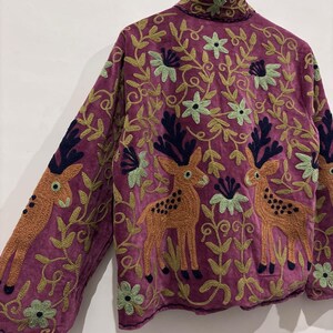 May include: A purple jacket with embroidered deer and floral patterns. The jacket features orange deer with black antlers and green and blue floral designs. The jacket is made of a soft, textured fabric.