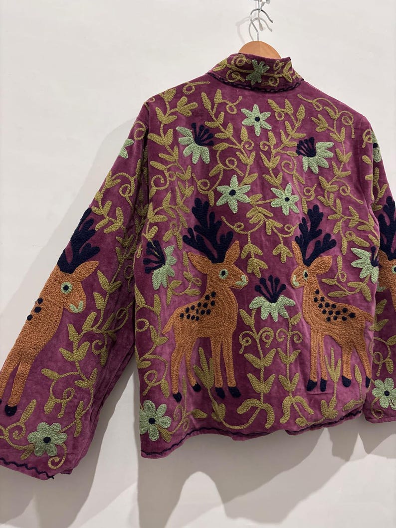May include: A purple jacket with embroidered deer and floral patterns. The jacket features orange deer with black antlers and green and blue floral designs. The jacket is made of a soft, textured fabric.