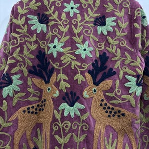 May include: A burgundy jacket with intricate embroidery. The design features two deer facing each other, surrounded by floral and leaf patterns in shades of green, brown, and navy. The jacket has a high collar.