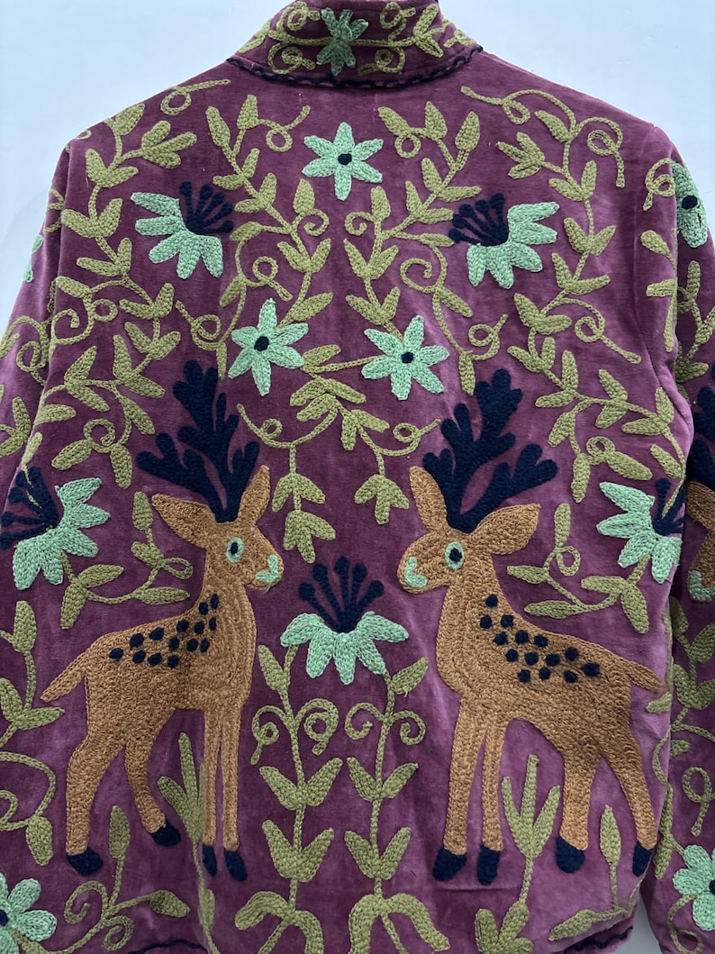 May include: A burgundy jacket with intricate embroidery. The design features two deer facing each other, surrounded by floral and leaf patterns in shades of green, brown, and navy. The jacket has a high collar.