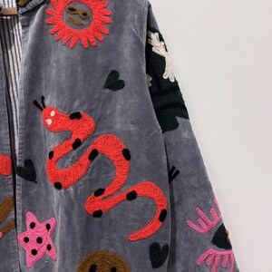 Puede incluir: Chaqueta gris con parches bordados coloridos. La chaqueta presenta un sol rojo, una serpiente roja con lunares negros, una estrella rosa, una cara sonriente marr&oacute;n y otros dise&ntilde;os caprichosos. La chaqueta tiene un forro a rayas y un cuello decorativo.