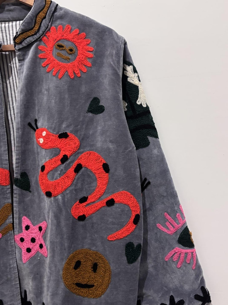 Puede incluir: Chaqueta gris con parches bordados coloridos. La chaqueta presenta un sol rojo, una serpiente roja con lunares negros, una estrella rosa, una cara sonriente marr&oacute;n y otros dise&ntilde;os caprichosos. La chaqueta tiene un forro a rayas y un cuello decorativo.