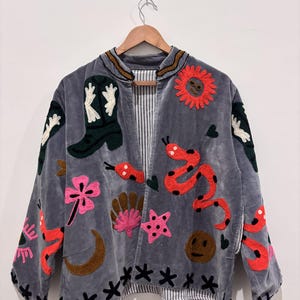 Puede incluir: Chaqueta de terciopelo gris con dise&ntilde;os bordados coloridos. Presenta una bota de vaquero, un sol, serpientes, flores y estrellas. La chaqueta tiene un forro a rayas y est&aacute; colgada en una percha de madera.