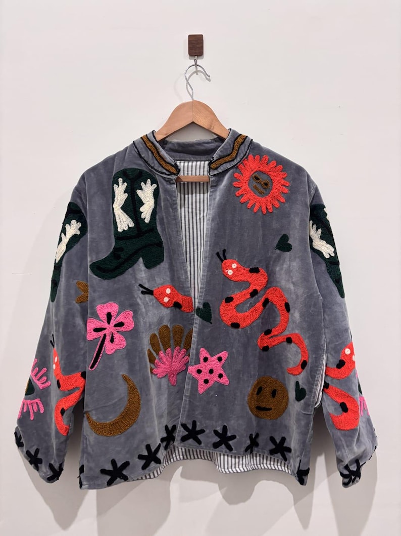 Puede incluir: Chaqueta de terciopelo gris con dise&ntilde;os bordados coloridos. Presenta una bota de vaquero, un sol, serpientes, flores y estrellas. La chaqueta tiene un forro a rayas y est&aacute; colgada en una percha de madera.