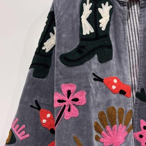 Puede incluir: Chaqueta de terciopelo gris con parches bordados. Los parches incluyen una bota de vaquero verde con detalles blancos, flores rosas y dise&ntilde;os de insectos rojos y negros. La chaqueta tiene un forro a rayas y un ribete marr&oacute;n y dorado.