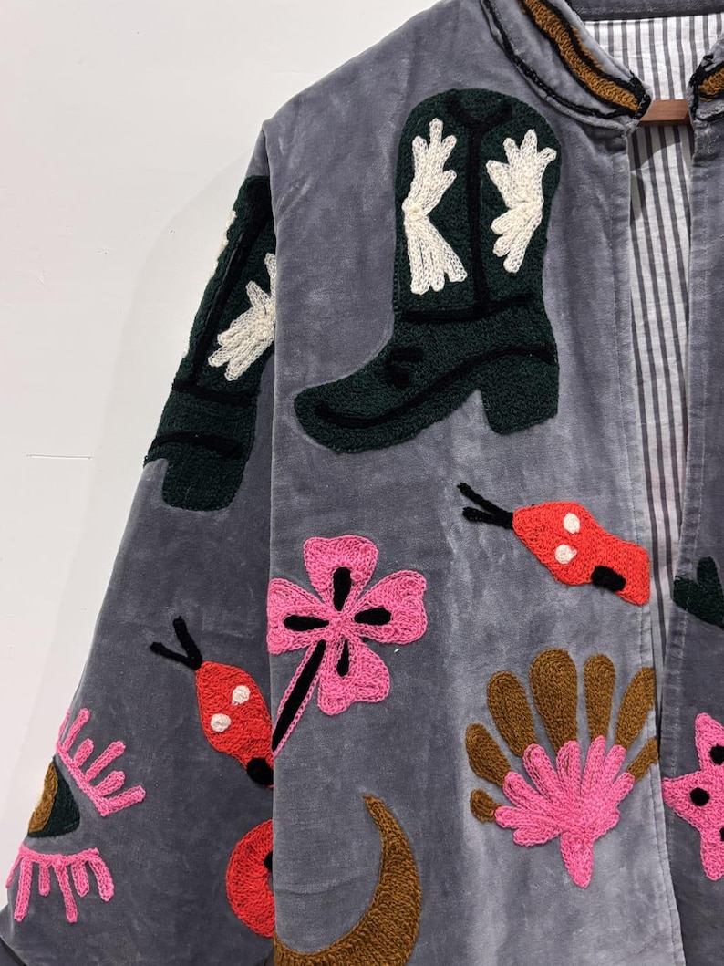 Puede incluir: Chaqueta de terciopelo gris con parches bordados. Los parches incluyen una bota de vaquero verde con detalles blancos, flores rosas y dise&ntilde;os de insectos rojos y negros. La chaqueta tiene un forro a rayas y un ribete marr&oacute;n y dorado.